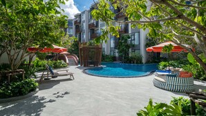 A pool area with lounge chairs and umbrellas, surrounded by greenery and a multi-story building. - Canvas Condo New 2BR Near Beach 111-80 (Choeng Thale)