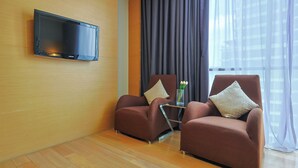 A modern room with two brown armchairs, a small table with a vase, a flat-screen TV mounted on the wall, and a large window with curtains. - Empire Suites Subang by Mana Mana (Subang Jaya)