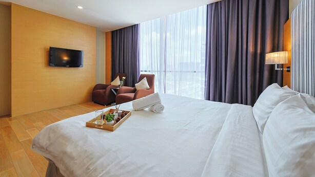 A hotel room with a bed, a TV mounted on the wall, a seating area with a chair and a table, and a large window with curtains. - Empire Suites Subang by Mana Mana (Subang Jaya)