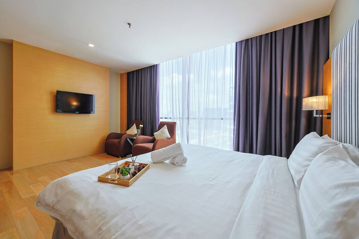 A hotel room with a bed, a TV mounted on the wall, a seating area with a chair and a table, and a large window with curtains.
