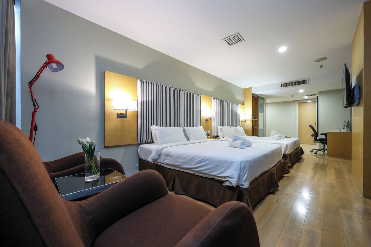 A hotel room with two beds, a desk, a chair, a TV, and a bathroom visible in the background.