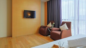 A hotel room with a wooden wall, a flat-screen TV, two armchairs, a bed, and a large window with sheer curtains. - Empire Suites Subang by Mana Mana (Subang Jaya)