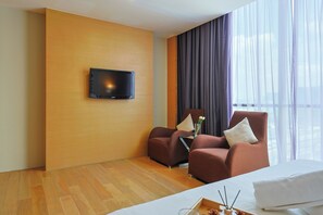 Habitación de hotel con pared de madera, televisor de pantalla plana, dos sillones, una cama y un ventanal con cortinas traslúcidas.