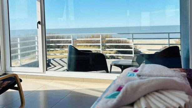 A balcony with a view of the beach, a bed with a blanket, and a chair.