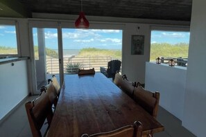 Ein geräumiger Essbereich mit einem langen Holztisch und Stühlen, großen Fenstern mit Blick auf den Strand und einer roten Pendelleuchte an der Decke.