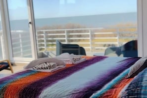 Ein Bett mit farbenfroher Decke und einem Paar Sneaker darauf, ein Fenster mit Meerblick und ein Stuhl im Hintergrund.