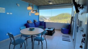 Ein modernes Zimmer mit blauer Wand, einem runden Tisch mit Stühlen, einer Couch mit blauen Kissen und einem großen Fenster mit Blick auf den Strand.