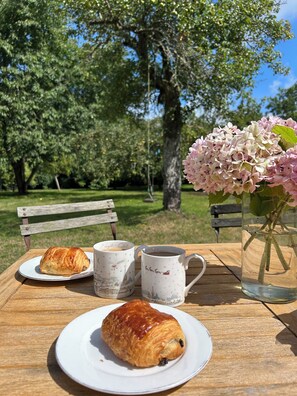 A picnic setup with a croissant, two mugs, and a vase of flowers on a wooden table in a park. - Our 17th century Barn converted into a relaxed, bohemian home. (Honfleur)