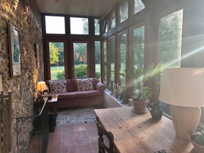 Une pièce avec un mur en pierre, une table en bois, un canapé agrémenté de coussins à motifs, une lampe et une vue sur la verdure à l’extérieur.