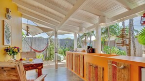 Un'area pranzo all'aperto coperta con tavolo e sedie in legno, un bancone bar con pensili in legno e vista su palme.