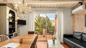 A dining area with a wooden table, chairs, and a chandelier. There is a black leather sofa and a small white table with a plant. A television is mounted on the wall, and a window provides a view of the outside. - Gràcia Loft + Terrace BBQ • AC & Fiber Wi-Fi (Barcelona)