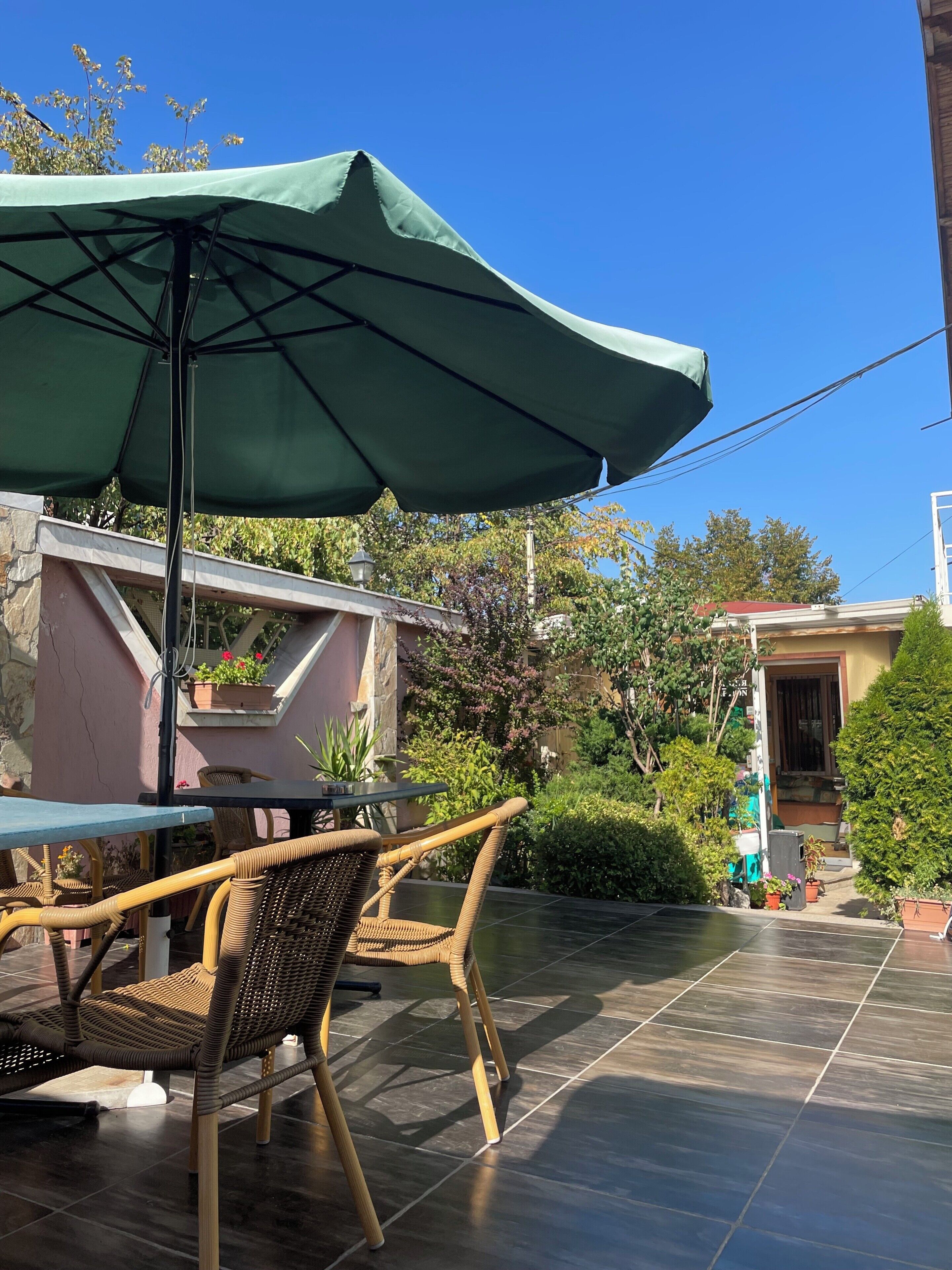An outdoor patio area with a table and chairs under a green umbrella, surrounded by potted plants and a stone wall.