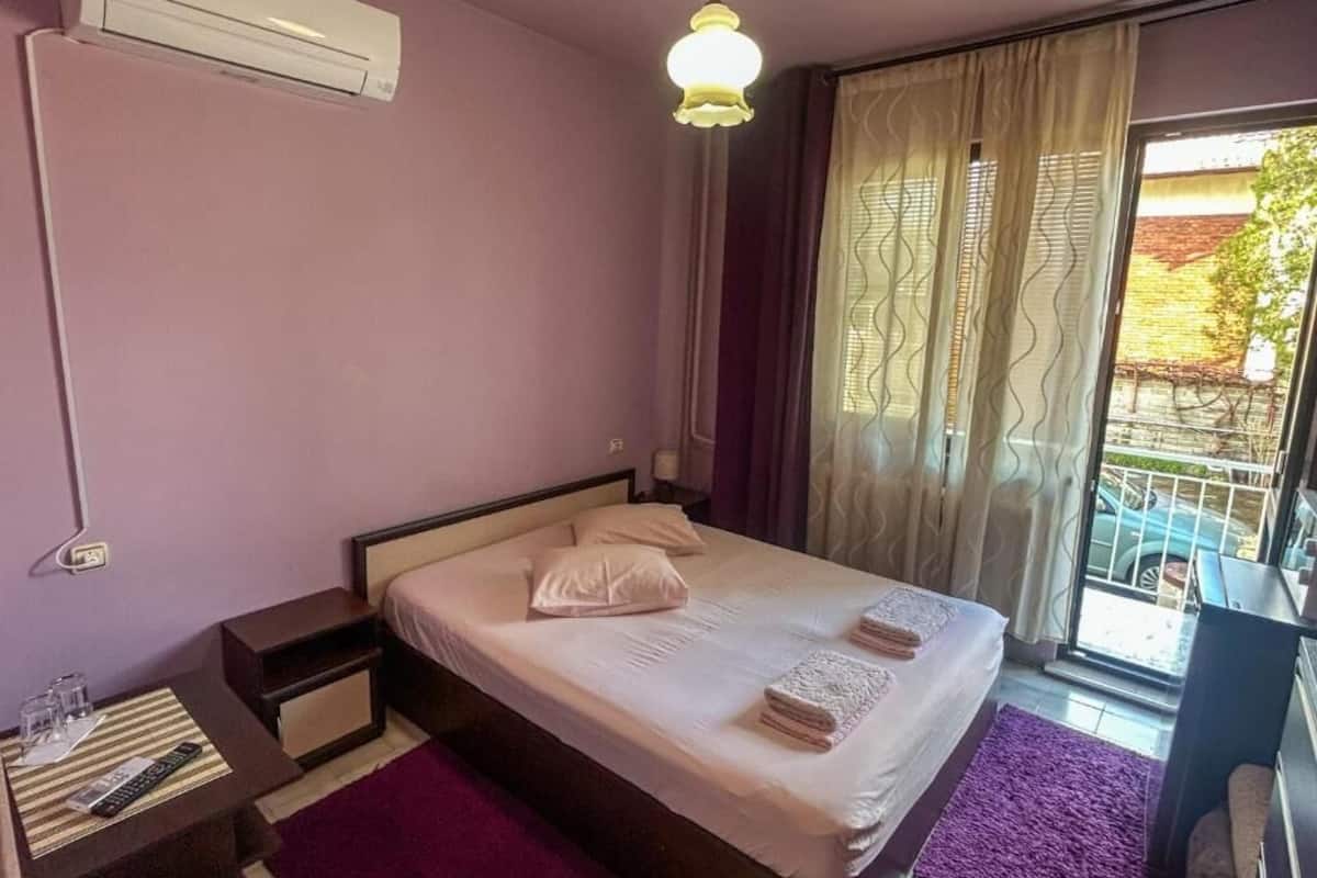 A hotel room with a bed, a nightstand, a window with curtains, and a balcony.
