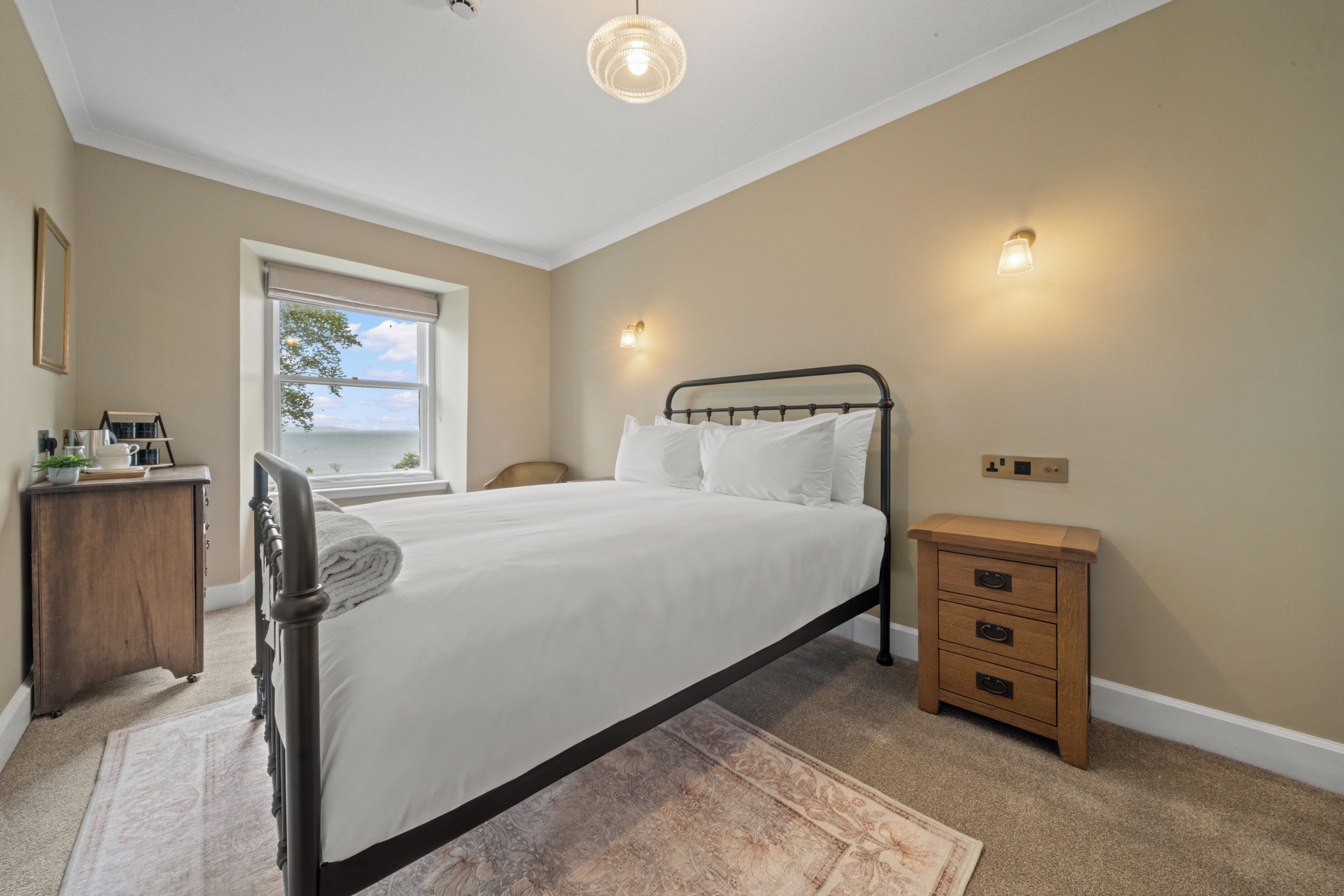 A bedroom with a bed, a nightstand, a dresser, a window with a view, and wall-mounted lights.