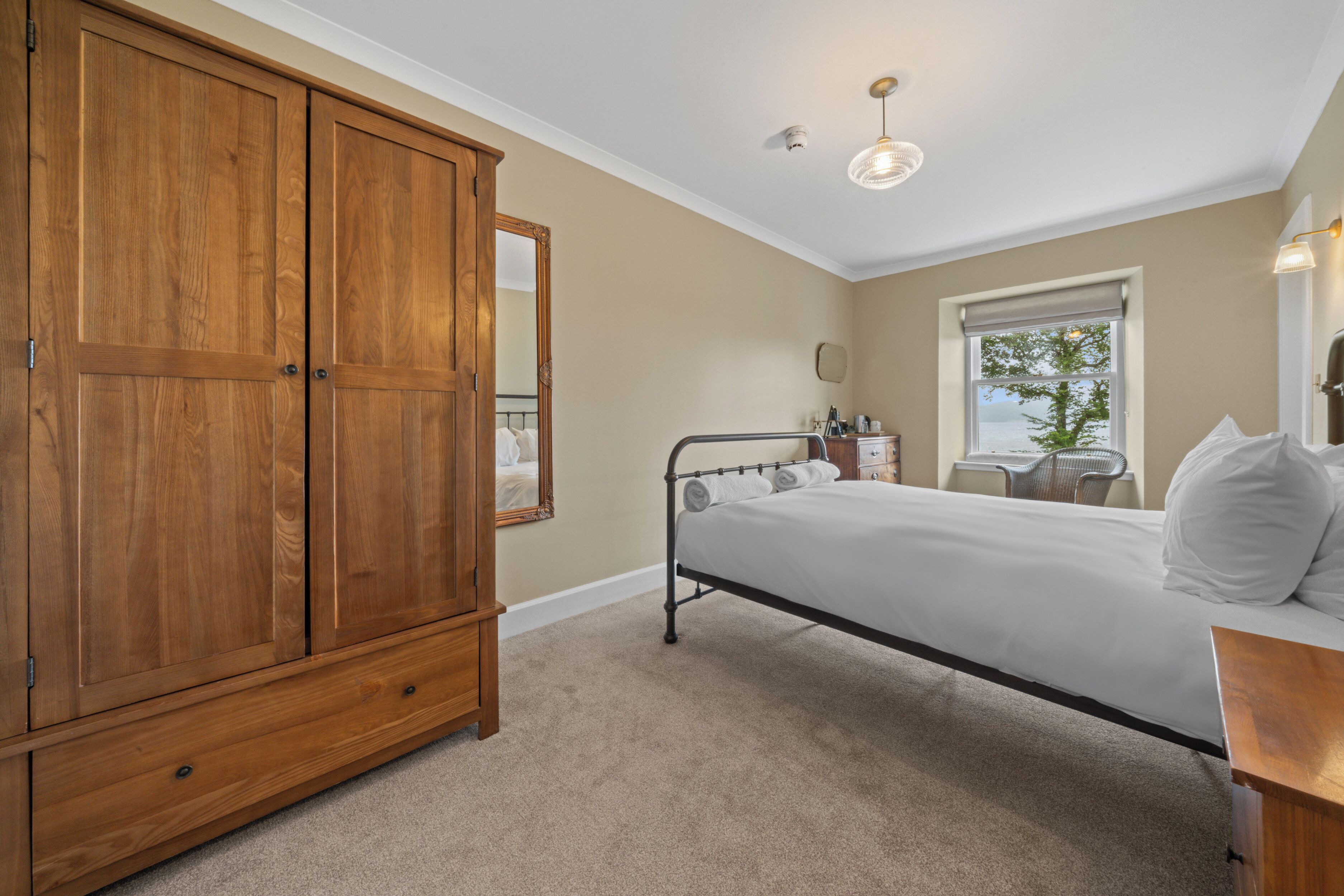 A bedroom with a large wooden wardrobe, a single bed with white bedding, a small bedside table, and a window with a view of trees.