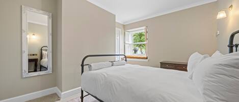 A bedroom with a bed, a nightstand, a dresser, a mirror, and a window with a view of greenery.