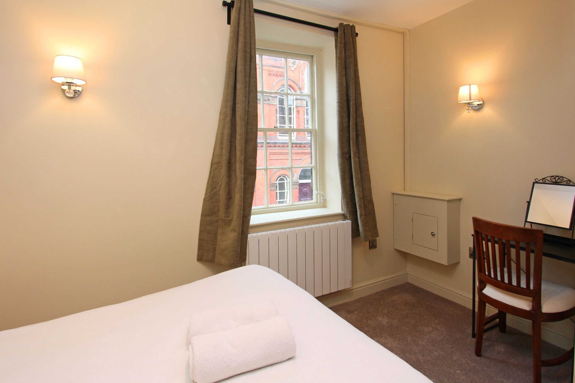 A bedroom with a bed, a chair, a desk, a window with curtains, and wall-mounted lights.