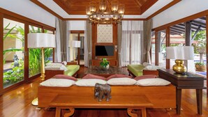 A spacious living room with a wooden ceiling, a large chandelier, a sofa set, a coffee table, and a television. - Royal Legacy Golf Villa (Rawai)