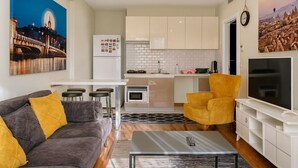 Ein kompakter Wohnbereich mit Sofa, Sesseln, einem Couchtisch, einem Fernseher, einer Küchenzeile inklusive Kühlschrank, Mikrowelle und Schränken sowie einem Stadtbild.