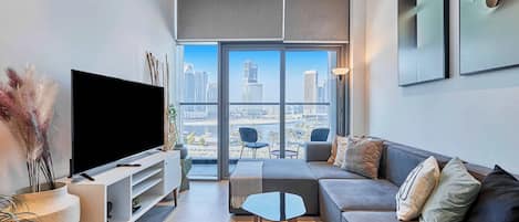 A modern living room with a flat-screen TV, a grey sofa, a small coffee table, and a large window offering a city view.