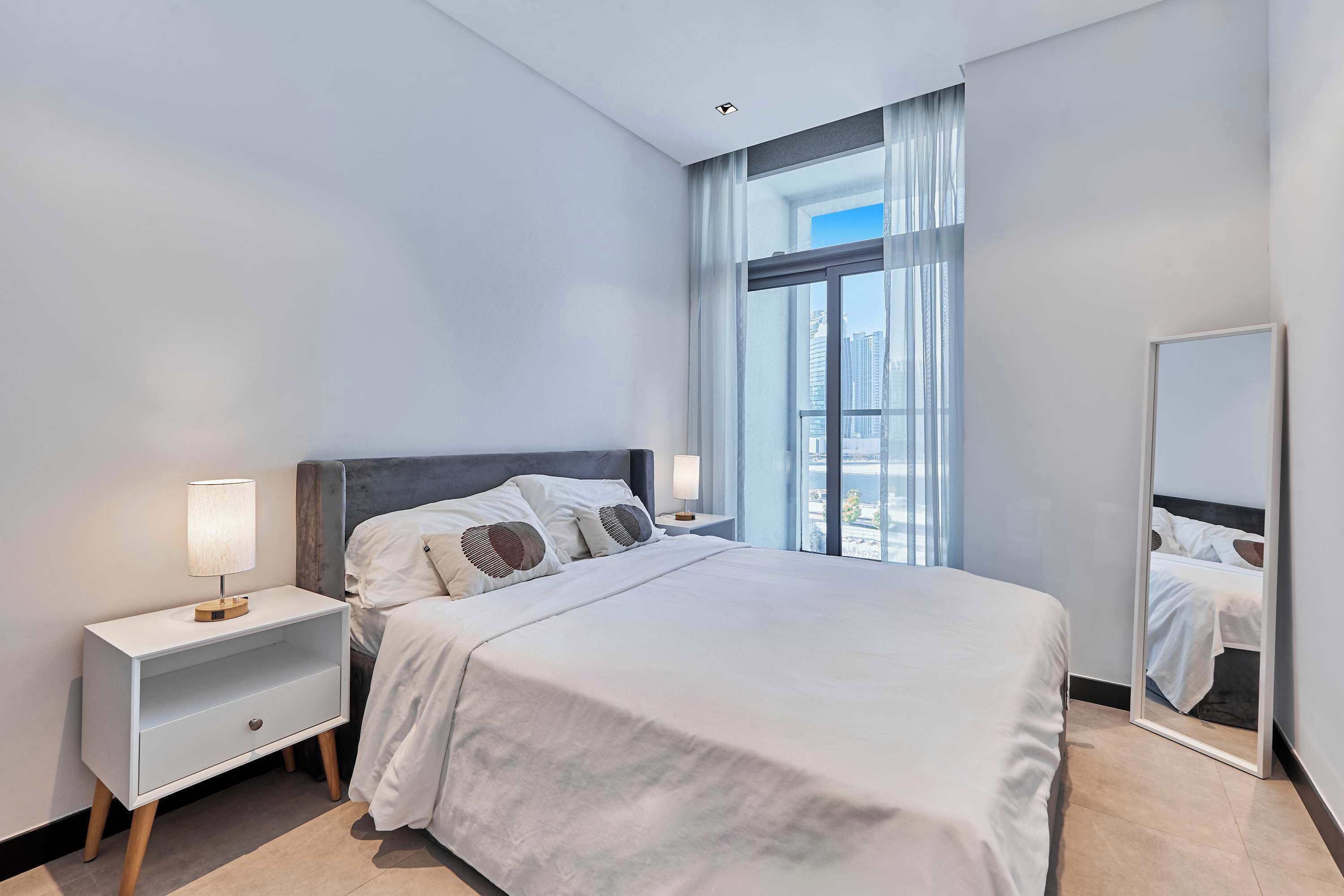 A modern hotel room with a large bed, a nightstand with a lamp, a mirror, and a view of the city through large windows.