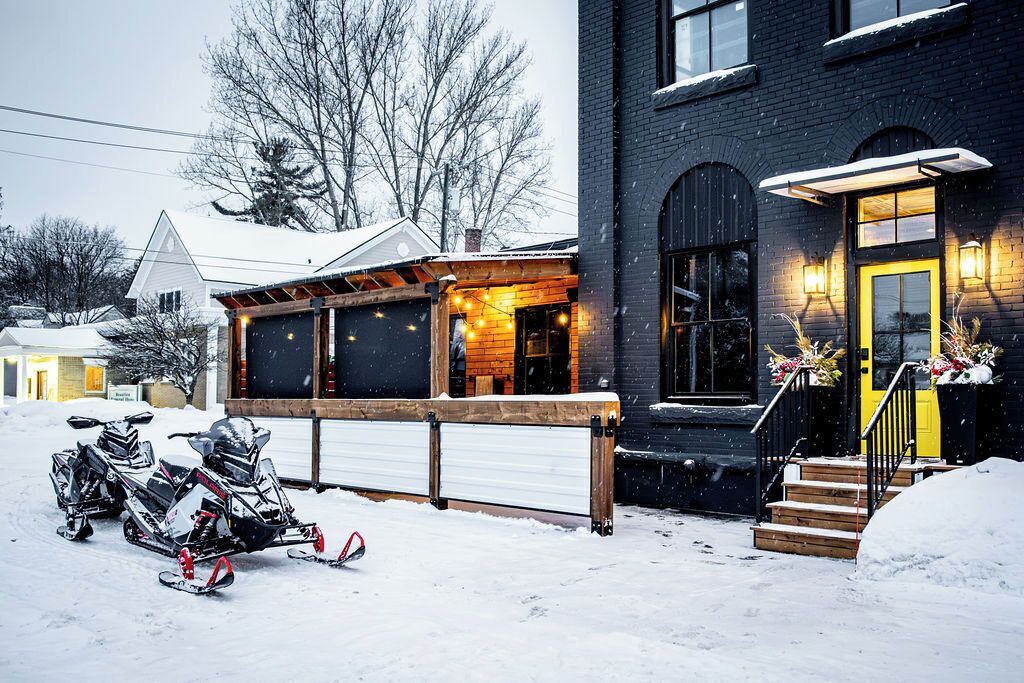 Et snedækket boligområde med et hus, der har en træveranda, en gul dør og en garage. Der er sne scootere parkeret foran.