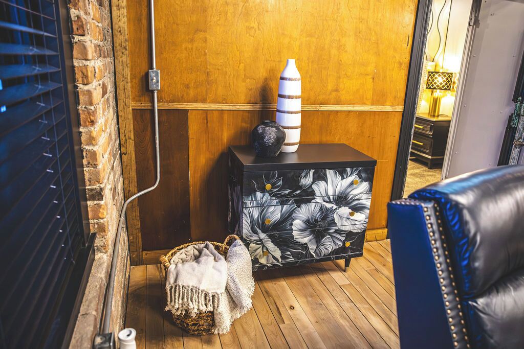 Une pièce avec un plancher en bois, une commode à motifs floraux noir et blanc, un fauteuil en cuir bleu et un mur en briques.