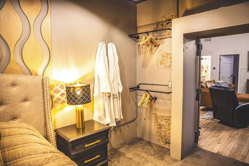 A hotel room with a tufted headboard, a bedside lamp, a wardrobe, a towel rack, and a mirror.