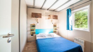 Una habitación estrecha con una cama, una ventana que da a zonas verdes y una estantería en la pared con libros.