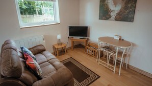 A living room with a brown sofa, a TV, a small table, and a window with a view of trees and a fence.