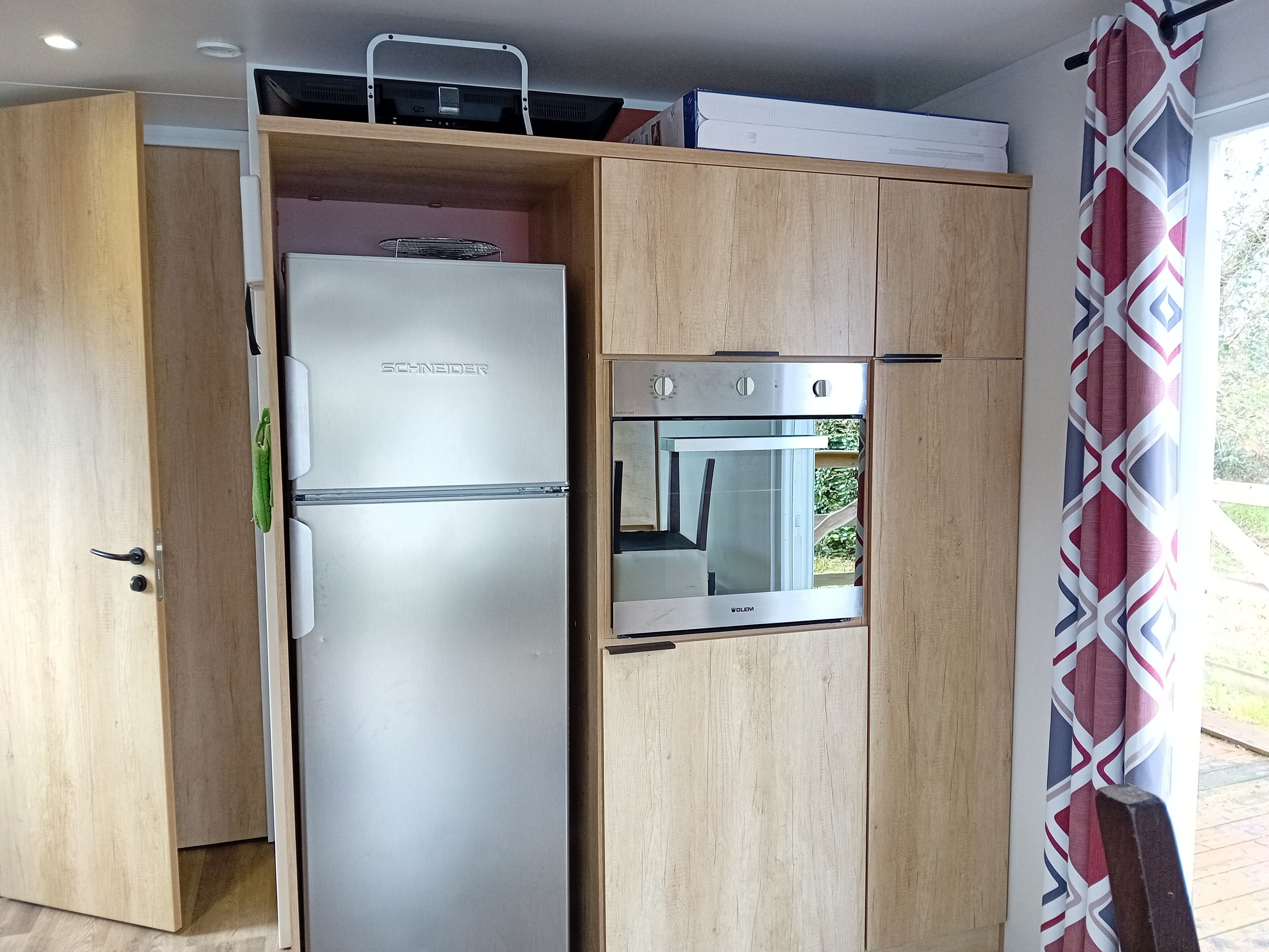 Une cuisine compacte dotée d’un réfrigérateur et d’un four encastrés, de meubles en bois et d’une fenêtre agrémentée de rideaux à motifs.