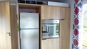 Eine kompakte Küche mit integriertem Kühlschrank und Backofen, Holzschränken und einem Fenster mit gemusterten Vorhängen.