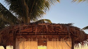 A beach hut with a thatched roof and white curtains, situated on a sandy beach with palm trees and the ocean in the background. - Surin Dreambeach View 4 (Thap Sakae)