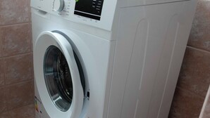 Une machine à laver compacte, de couleur blanche, à chargement frontal et dotée d’un panneau de commande numérique, située dans une salle de bain carrelée de carreaux beiges.