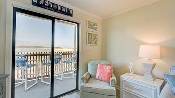 A room with a sliding glass door leading to a balcony with chairs and a view of the beach. There is a chair with a pink pillow, a bedside table with a lamp, and a framed map on the wall.