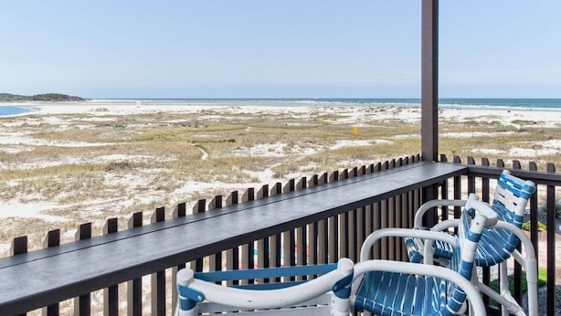 Blick auf einen Strand von einer überdachten Terrasse mit Holzgeländer, mit Sandlandschaft und blauem Himmel.