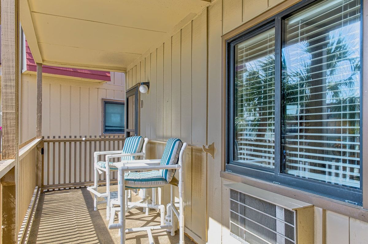 A balcony with two white chairs and a table, a window with blinds, and a door with a light fixture.