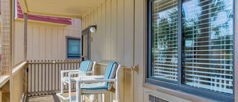 A balcony with two white chairs and a table, a window with blinds, and a door with a light fixture.
