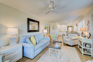 A living room with a blue sofa, a coffee table, and a white lamp. There is a ceiling fan and a kitchen in the background. - 2BR/2B Oceanfront & Inlet View Gated w/pool (North Myrtle Beach)
