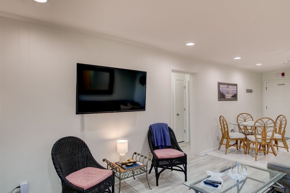 A hotel room with a flat-screen TV mounted on the wall, a dining area with a table and chairs, and a seating area with two chairs.