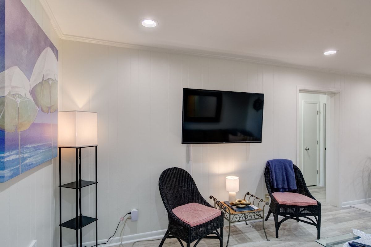 A living room with a flat-screen TV mounted on the wall, two wicker chairs with cushions, a floor lamp, and a painting on the wall.