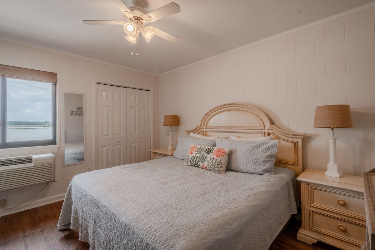 A bedroom with a bed, bedside lamps, a ceiling fan, a window, and a door.