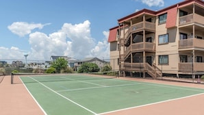 En tennisbana med nät, omgiven av ett staket. Det finns fler våningar höga byggnader med balkonger och en klar himmel.