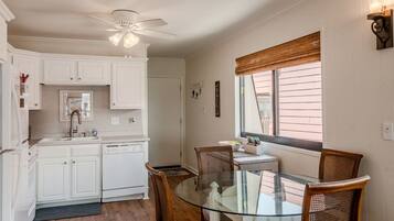 A kitchen with white cabinets, a glass dining table, wicker chairs, a ceiling fan, and a window with blinds.