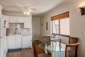 Une cuisine avec des meubles blancs, une table à manger en verre, des chaises en osier, un ventilateur de plafond et une fenêtre avec des stores.