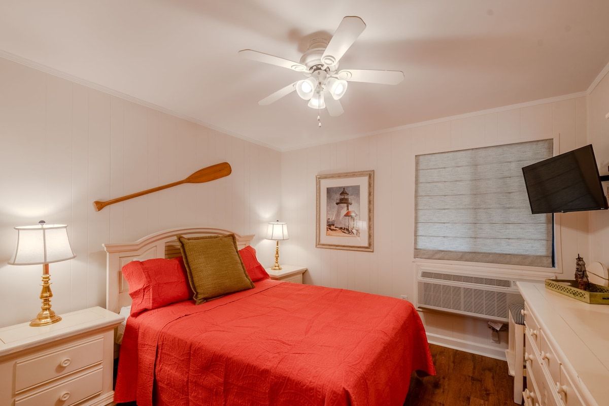Une chambre à coucher avec un lit, un ventilateur de plafond, une télévision et une fenêtre avec des stores.