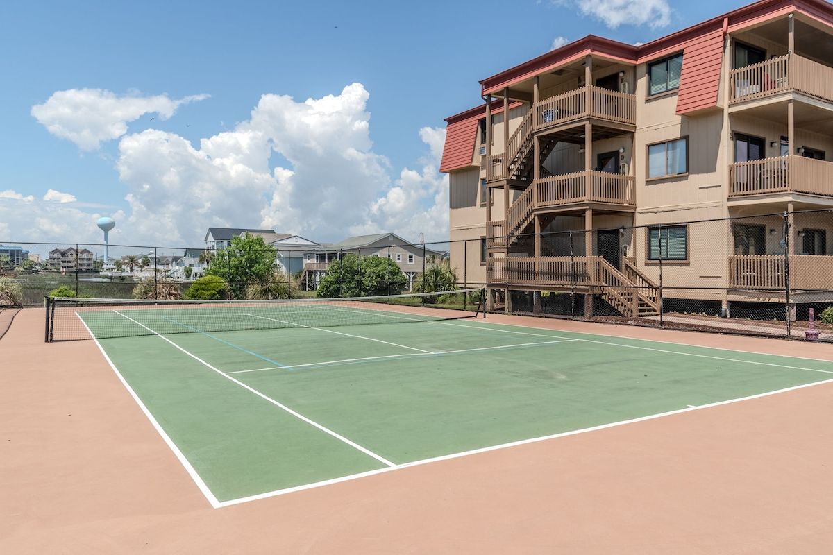 Un court de tennis avec un filet, entouré d’une clôture, et situé à côté d’un immeuble à plusieurs étages doté de balcons.