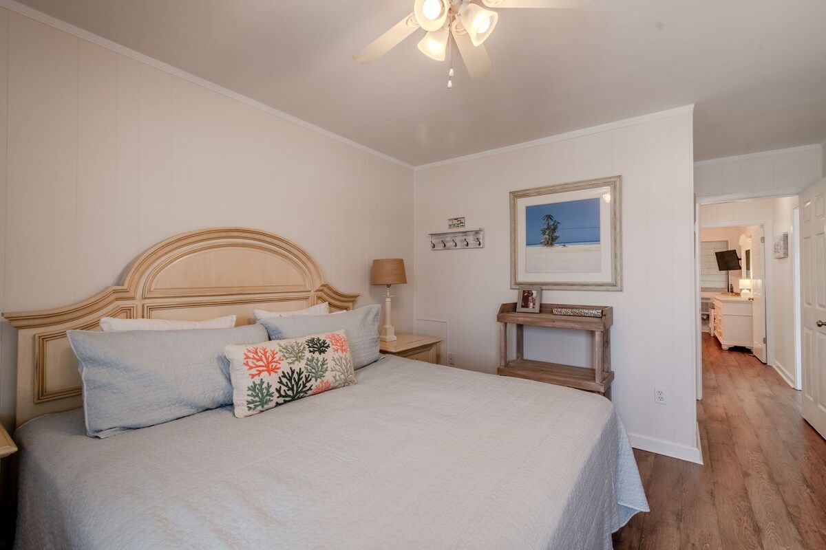 A bedroom with a bed, a nightstand, a ceiling fan, a framed picture, and a door leading to another room.