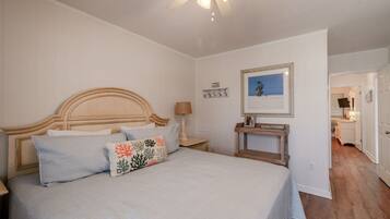 A bedroom with a bed, a nightstand, a ceiling fan, a framed picture, and a door leading to another room.