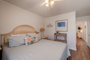 Une chambre à coucher comprenant un lit, une table de chevet, un ventilateur de plafond, un tableau encadré et une porte donnant sur une autre pièce.